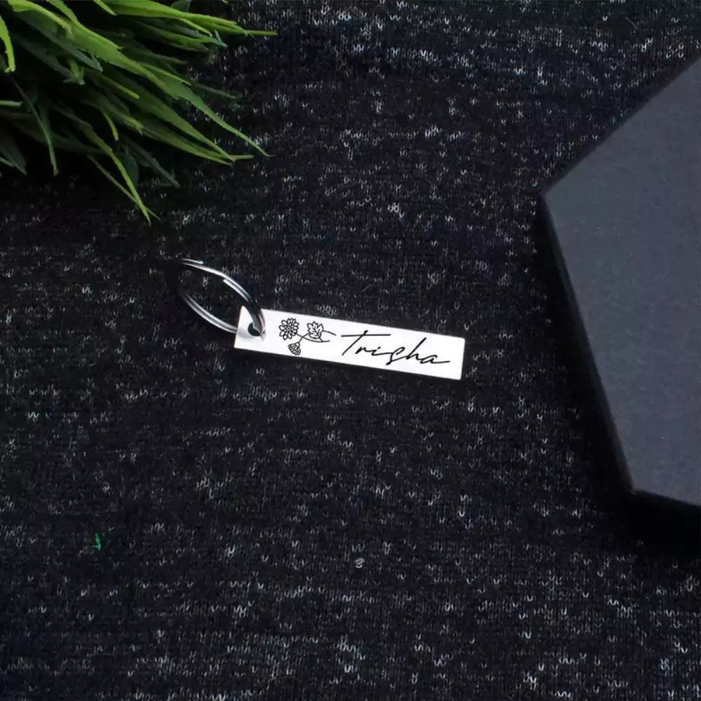 A customized birthday keychain with a flower charm and the name 'Thysha' engraved on it, placed on a textured surface next to a black box and some greenery.