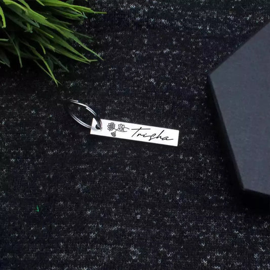 A customized birthday keychain with a flower charm and the name 'Thysha' engraved on it, placed on a textured surface next to a black box and some greenery.