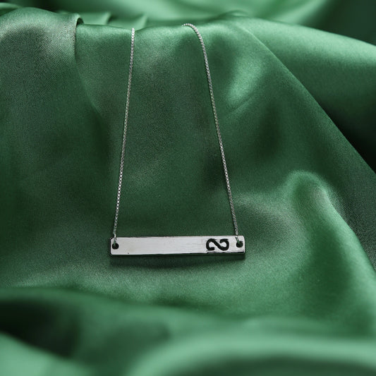 A silver bar necklace with a hand-stamped symbol pendant, displayed on draped green satin fabric.